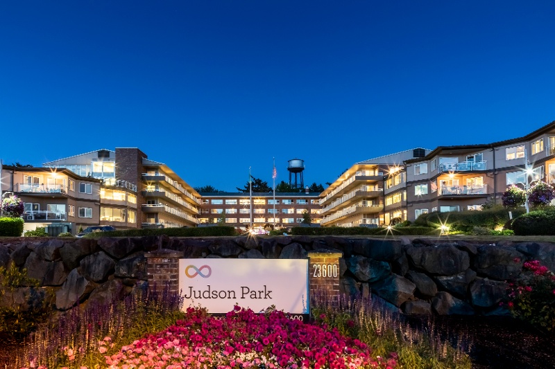 A condo building with illuminated windows. A sign surrounded by flowers reads 